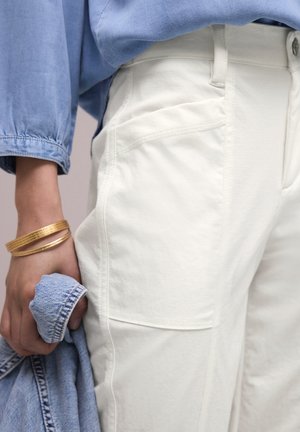 Personne portant un pantalon blanc et une chemise en jean bleue, tenant une veste en jean bleue, avec des bracelets en or au poignet.
