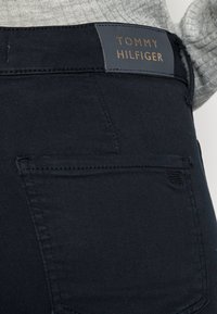 Jeans de mezclilla azul oscuro con un bolsillo trasero visible. Presenta una etiqueta de parche de cuero en navy con texto dorado que dice "TOMMY HILFIGER."