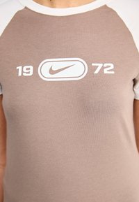Rudas medvilninis marškinėlis su baltais raglano tipo rankovėmis, turintis spausdintą Nike logotipą ir skaičius "19" bei "72" balta spalva. Lygus paviršius.