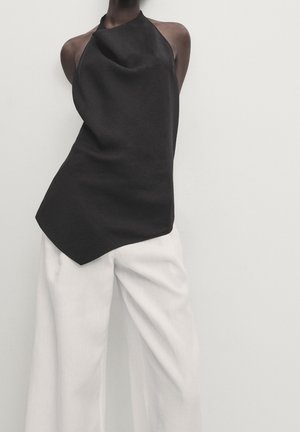 Persona che indossa un top asimmetrico nero senza maniche a collo halter abbinato a pantaloni bianchi larghi e ampi su sfondo semplice.