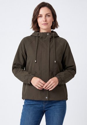 Donna che indossa una giacca con cappuccio verde oliva con coulisse e jeans blu, in piedi davanti a uno sfondo chiaro e semplice.