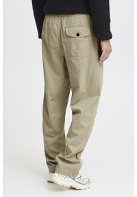 Pantalon en coton kaki avec une ceinture élastique, un design à jambe droite et une poche arrière ornée d'un bouton noir. Porté avec des chaussures de couleur claire.