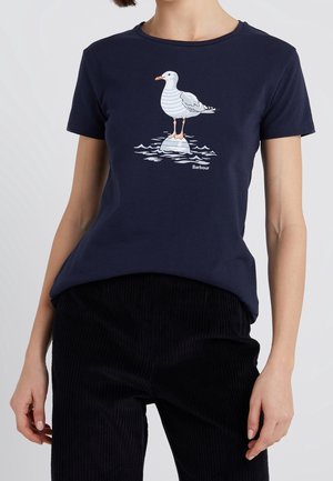 Kobieta w granatowej koszulce z grafiką mewy stojącej na skale w wodzie oraz w czarnych spodniach sztruksowych.