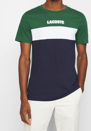 Hombre vistiendo una camiseta Lacoste color block en verde, blanco y azul marino con mangas cortas, combinada con pantalones blancos, de pie con una mano en el bolsillo.