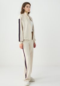 Beige Trackjacke und -hose aus weichem Stoff, mit einem hohen Kragen und kontrastierenden vertikalen Streifen in Lila und Marineblau.