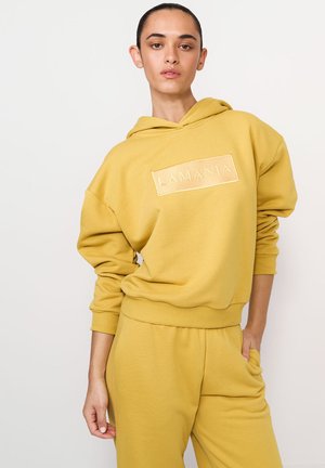 Żółta bluza z kapturem z teksturowanym logo "LAMANIA", z przednią kieszenią, ściągaczami przy rękawach i u dołu. W komplecie ze spodniami dresowymi w tym samym odcieniu żółci.