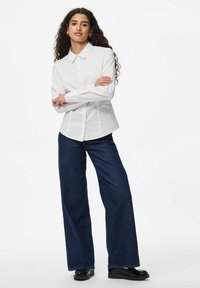 Camicia bianca con colletto, maniche lunghe e vita aderente, abbinata a jeans a gamba larga blu scuro e scarpe con plateau nere.