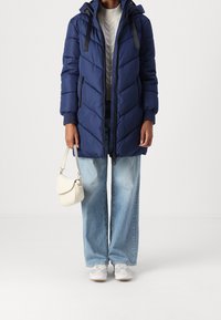 Granatowa kurtka puchowa z kapturem, z przeszyciami w kształcie jodełki, kieszeniami na zamek i ściągaczami przy mankietach. Połączona z jasnoniebieskimi jeansami i białymi trampkami.