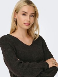 Tamni pleteni džemper s V-izrezom, blago opuštenog kroja i rebraste teksture. Model prikazuje naušnice u obliku obruča i prsten, s dugom plavom kosom.