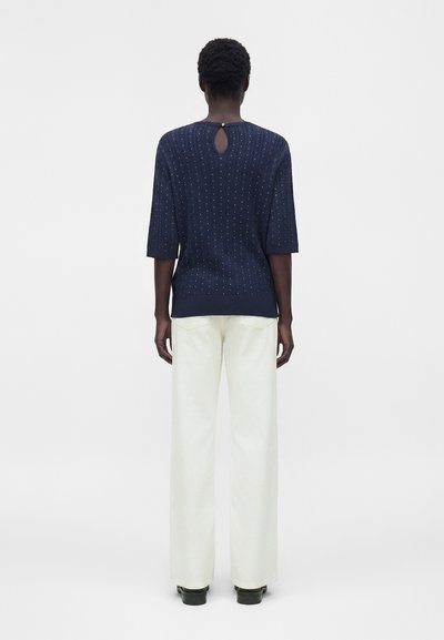 Άτομο που στέκεται με την πλάτη γυρισμένη, φοράει μπλε σκούφο πουλόβερ με μικρές κουκίδες, λευκό παντελόνι με φαρδιά μπατζάκια και μαύρα παπούτσια.