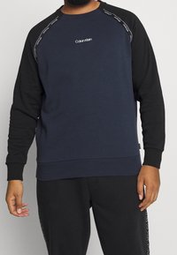 Námořnicky modrá mikina s černými raglánovými rukávy, žebrovanými manžetami a lemem. Na prsou je vyšitý bílý nápis "Calvin Klein".