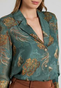 Blusa verde con estampado, con bordado floral metálico, cuello chal y cierre de botones, hecha de tejido ligero y texturizado.