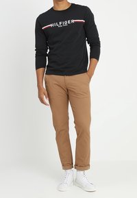 Mężczyzna w czarnej koszuli z długim rękawem Hilfiger, w beżowych spodniami podwiniętymi, w białych sneakersach z wysoką cholewką, stoi z jedną ręką w kieszeni na białym tle.