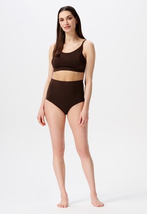 Vrouw die blote voeten staat, gekleed in een bruin hooggesneden broekje en een bijpassend mouwloos crop top voor een witte achtergrond.