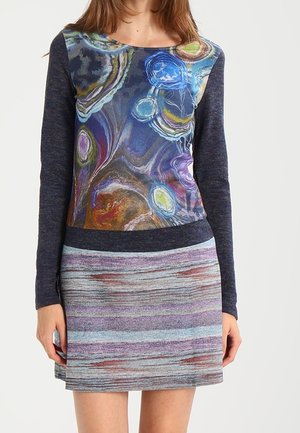 Frau, die ein langärmliges Kleid mit einem bunten abstrakten Wirbelmuster am Oberteil und einem horizontal gestreiften Rock in gedämpften Tönen trägt.