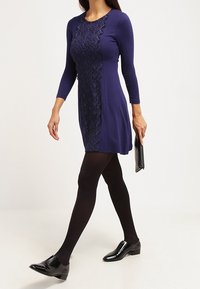 Marinblå klänning med långa ärmar, prydd med spetsdetaljer längs mitten. Bärs med svarta strumpbyxor och blanka svarta loafers. Clutch i handen.