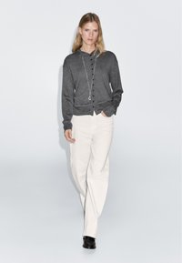 Cardigan gris à boutons avec manches longues, associé à un pantalon large blanc et des chaussures noires ; texture lisse et coupe décontractée.