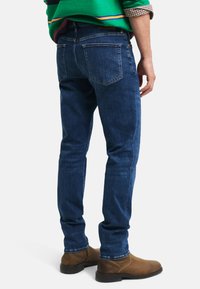 Jeans de mezclilla en azul oscuro, corte ajustado; cuentan con dos bolsillos traseros, costuras amarillas y costuras laterales a lo largo de las piernas. Combinados con botas marrones.