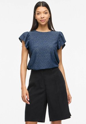 Femme portant un chemisier bleu marine à manches courtes ajouré et un short noir à la coupe ajustée, longueur genou, debout sur fond blanc.