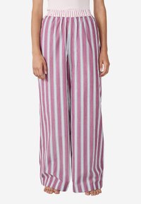 Kobieta w luźnych, szerokich spodniach z pionowymi różowymi, białymi i niebieskimi paskami, stojąca boso na białym tle.