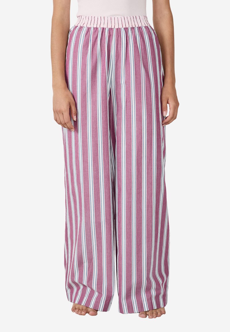 Kobieta w luźnych, szerokich spodniach z pionowymi różowymi, białymi i niebieskimi paskami, stojąca boso na białym tle.