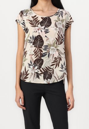 Femme portant un chemisier beige à manches courtes avec un imprimé feuilles et fleurs tropicales mélangées marron foncé et vert, assorti à un pantalon noir.