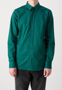 Chemise boutonner en teal avec un col, des manches longues et des boutons noirs ; présente une texture lisse et une coupe ajustée. Portée avec un pantalon sombre.