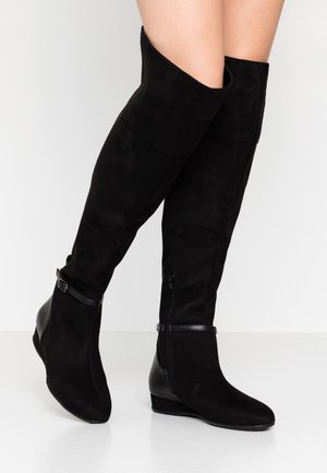 Bottes en daim noir montant jusqu'aux genoux avec fermeture éclair latérale et sangle à la cheville, portées sur des jambes nues, debout sur un sol blanc devant un fond clair.