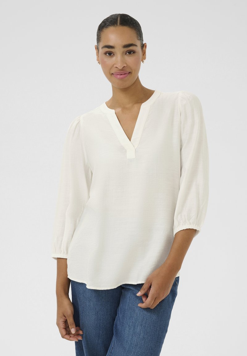 Blouse blanche légère avec un col en V et des manches bouffantes trois-quarts. Fabriquée en tissu texturé, elle présente des poignets élastiques et une coupe décontractée.