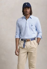 Világoskék, hosszú ujjú pólóing zsebbel, bézs rakott nadrág és csíkozott öv. Sötétkék sapka logóval, óra a csuklón.