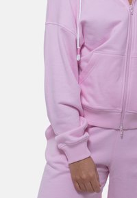 Lichtroze rits hoodie van zacht materiaal, met een rits aan de voorkant, een trekkoord en een geribde zoom en manchetten. Soepele textuur zonder patronen.