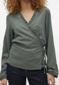 Grön blus med våfflad detalj längs halsringningen och ärmsluten. Tillverkad av textil med struktur och har en knytning på sidan.