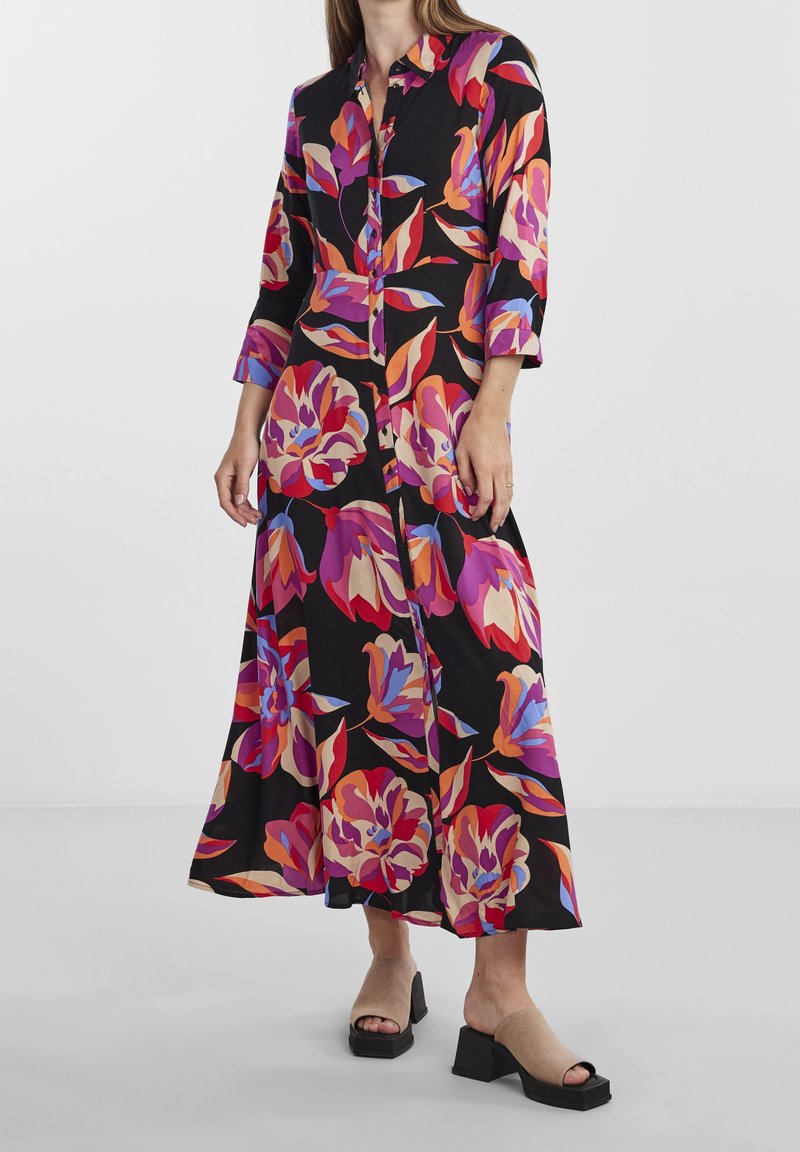 Femme portant une longue robe noire à imprimé floral large rouge, violet et orange avec des manches trois-quarts, associée à des talons blocs beiges à bout ouvert.