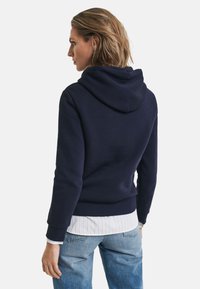 Marineblå hettegenser laget av myk stoff, med en romslig hette og ribbestrikkede mansjetter, brukt over en hvit stripete skjorte og lyse denimjeans.