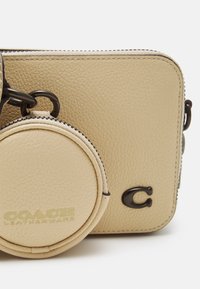 Beige läderaxelväska med kornig textur, utrustad med en avtagbar rund myntväska och präglad "COACH"-logotyp på framsidan. Svarta metallaccenter.