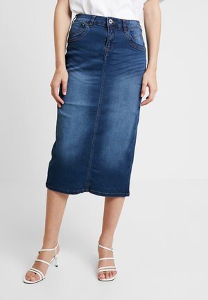 Frau trägt einen blauen Denim-Midirock und weiße, verzierte Sandaletten mit Absatz, steht vor einem schlichten hellen Hintergrund.