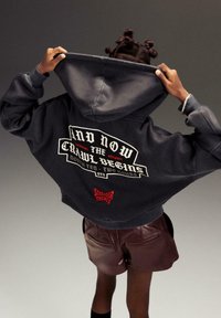 Čierna skrátená mikina s kapucňou a grafickým textom na chrbte. Obsahuje logo "Stranger Things" v červenej farbe. Materiál vyzerá mäkko a má uvoľnený strih.