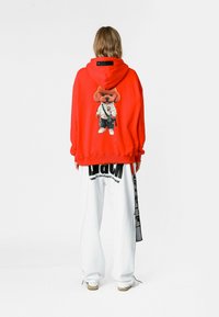 Sudadera roja con capucha que presenta un gráfico de un perro de dibujos animados en la parte posterior. Combinada con pantalones holgados blancos que exhiben texto negro y correas.