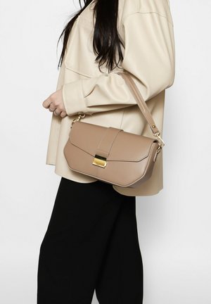 Woman wearing beige jacket and black pants holding tan leather handbag with gold clasp over shoulder.