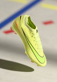 Botín de fútbol amarillo brillante con un upper texturizado, acentos negros y un swoosh verde neón. Presenta un diseño de cordones y una suela con tacos rojos.