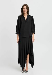 Black pleated blouse with a v-neck, balloon sleeves, paired with an asymmetrical black skirt. Model also wears silver pointed-toe heels.
