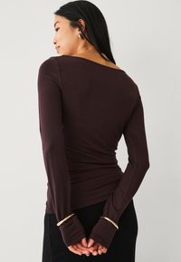 Haut à manches longues en tissu extensible bordeaux riche, avec un décolleté arrondi et des poignets avec trou pour le pouce uniques, au design élégant et ajusté.