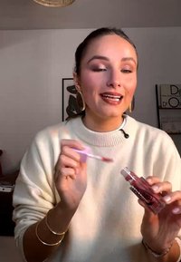 Ung kvinde iført guldringe og cremefarvet sweater holder og påfører lyserød læbepomade indendørs.