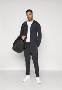 Zwarte sportjack en -broek met logo-accenten. Witte t-shirt eronder. Zwarte sporttas met gestructureerde afwerking. Witte sneakers.