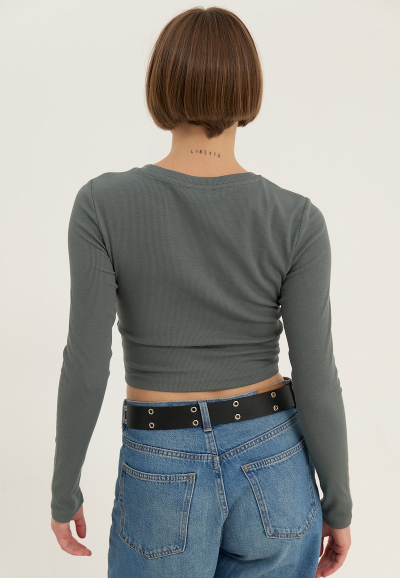Camisa de manga larga y cropped en tela de canalé verde apagado, combinada con jeans de denim azul y un cinturón negro con detalles de ojales.