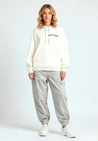 Felpa cremosa con testo rosso e nero, abbinata a pantaloni cargo grigi chiari, con polsini elasticizzati e tasche laterali, indossata con sneakers bianche.