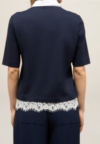 Námořnická modrá blůza s krátkým rukávem a bílým límečkem, se krajkovým lemem na spodním okraji, s rovnou siluetou a hladkou texturou látky.