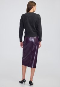 Vrouw in een donkergrijze trui, glanzende paarse leren rok tot de knie, zwarte kitten heels en gouden ringoorbellen, staande met de rug naar de kijker.