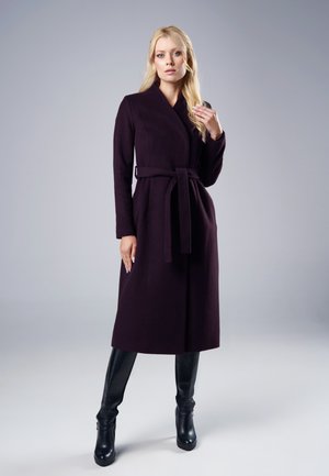 Blondynka w długim ciemnofioletowym płaszczu z paskiem i czarnych kozakach za kolano, stojąca na tle jasnoszarej, jednolitej ściany.