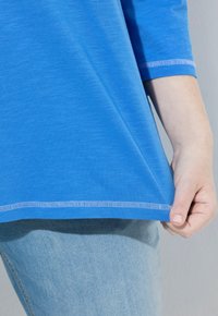 Sininen puuvillapaita, jossa on kolme neljänneksen hihavaa, kaareva helmus ja kontrastiväriset valkoiset tikkausompeleet, yhdistettynä vaaleansinisiin farkkuihin.